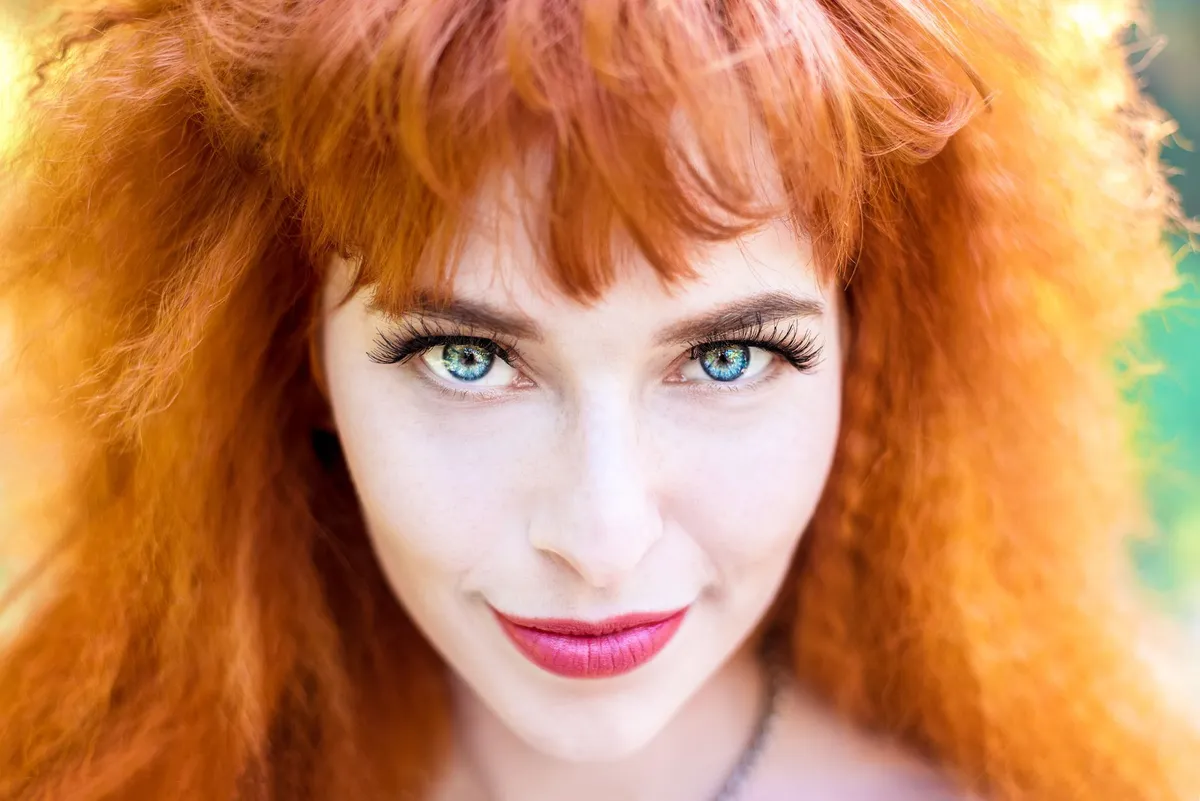 Femme rousse aux cheveux longs et ondulés, souriante, avec des cils longs et des lèvres rouges.